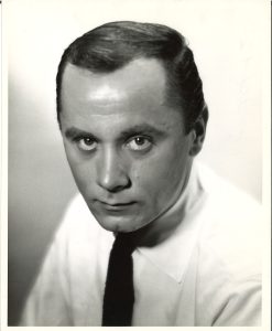 Professional black and white head shot of an approximately 40 year old white male looking upwards at the camera and wearing a light colored shirt and dark tie.